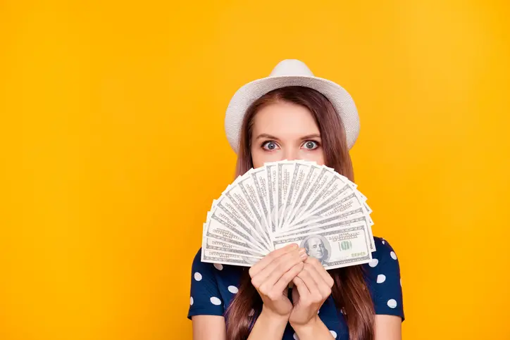 Portrait of pretty, charming, girl holding in hand a lot of money, close with fan half face, looking out with eyes, having white hat on head, isolated on yellow background Portrait of pretty, charming, girl holding in hand a lot of money, close with fan half face, looking out with eyes, having white hat on head, isolated on yellow background